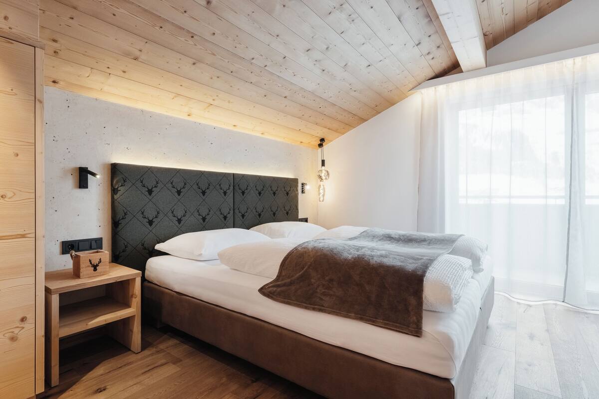 Quarto com teto de madeira, cama com roupa de cama branca, cabeceira com padrão geométrico e mesa de cabeceira com caixa de madeira.