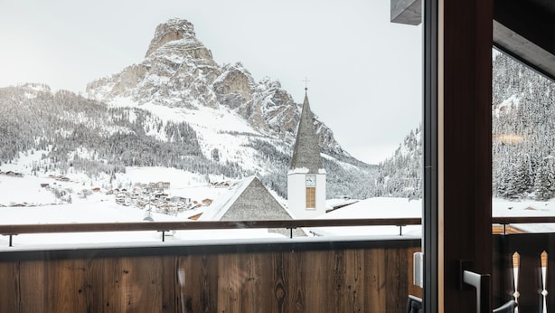 A wooden balcony with a view of a snow-covered mountain and a church. - Villa Trieste (Corvara in Badia)