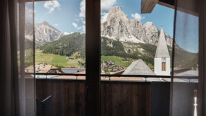 A view from a balcony with a wooden railing, overlooking a mountainous landscape, a church, and a small village. - Villa Trieste (Corvara in Badia)