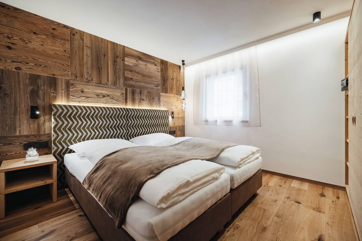 Quarto com cabeceira de madeira, cama com roupa de cama branca, mesa de cabeceira e janela com cortinas finas.