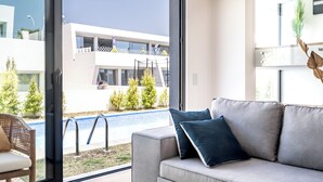 Un salon moderne avec un canapé gris, un fauteuil en osier et une vue sur une piscine et des bâtiments à l’extérieur.