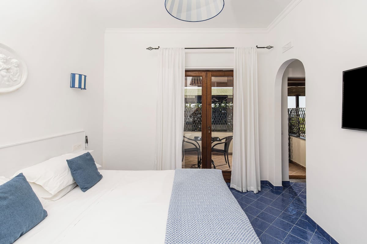 A bedroom with a bed, white walls, a blue patterned bedspread, and a view of the balcony.