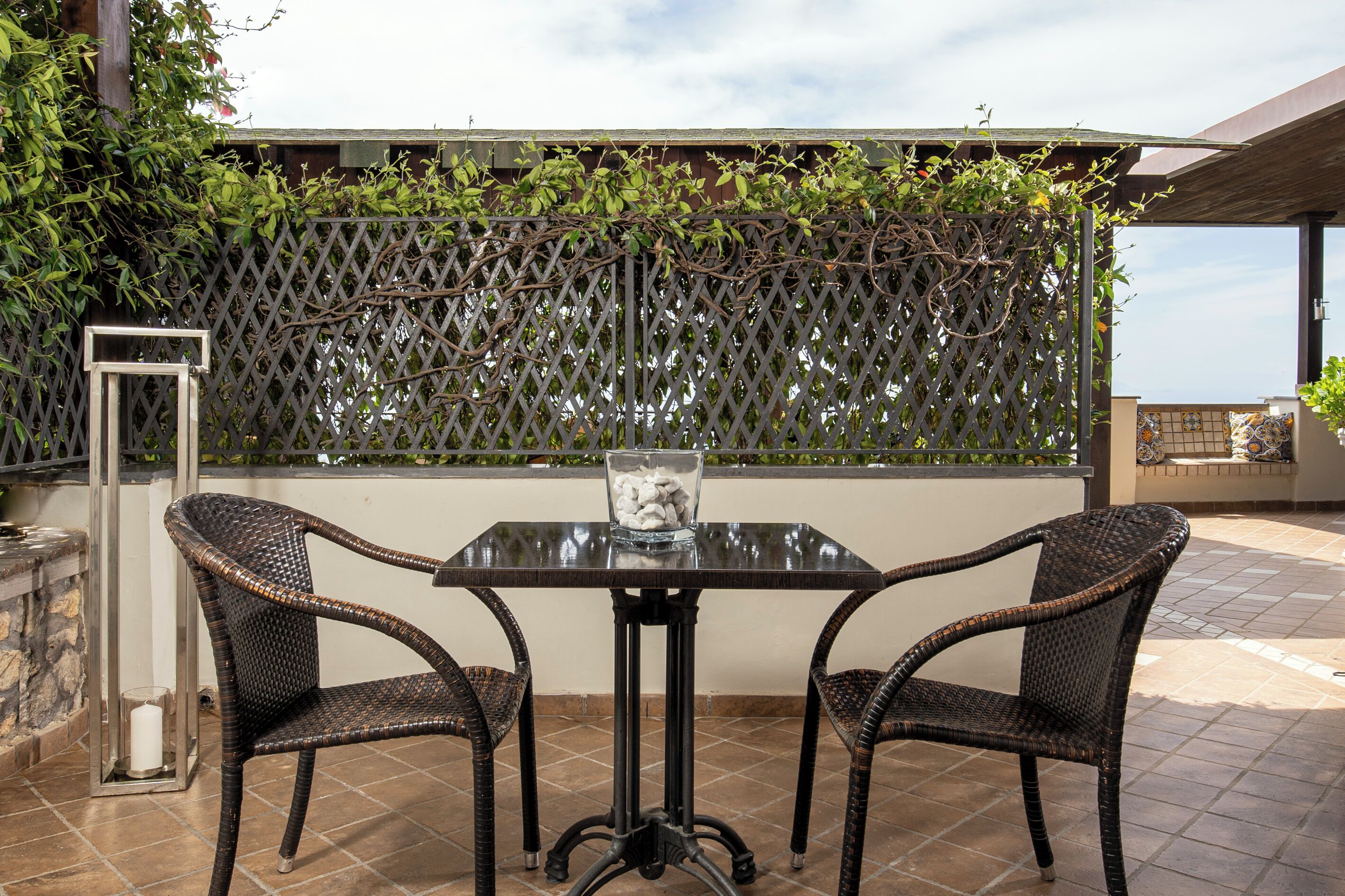 Un patio al aire libre con una mesa de vidrio, dos sillas de mimbre y un florero de vidrio decorativo.