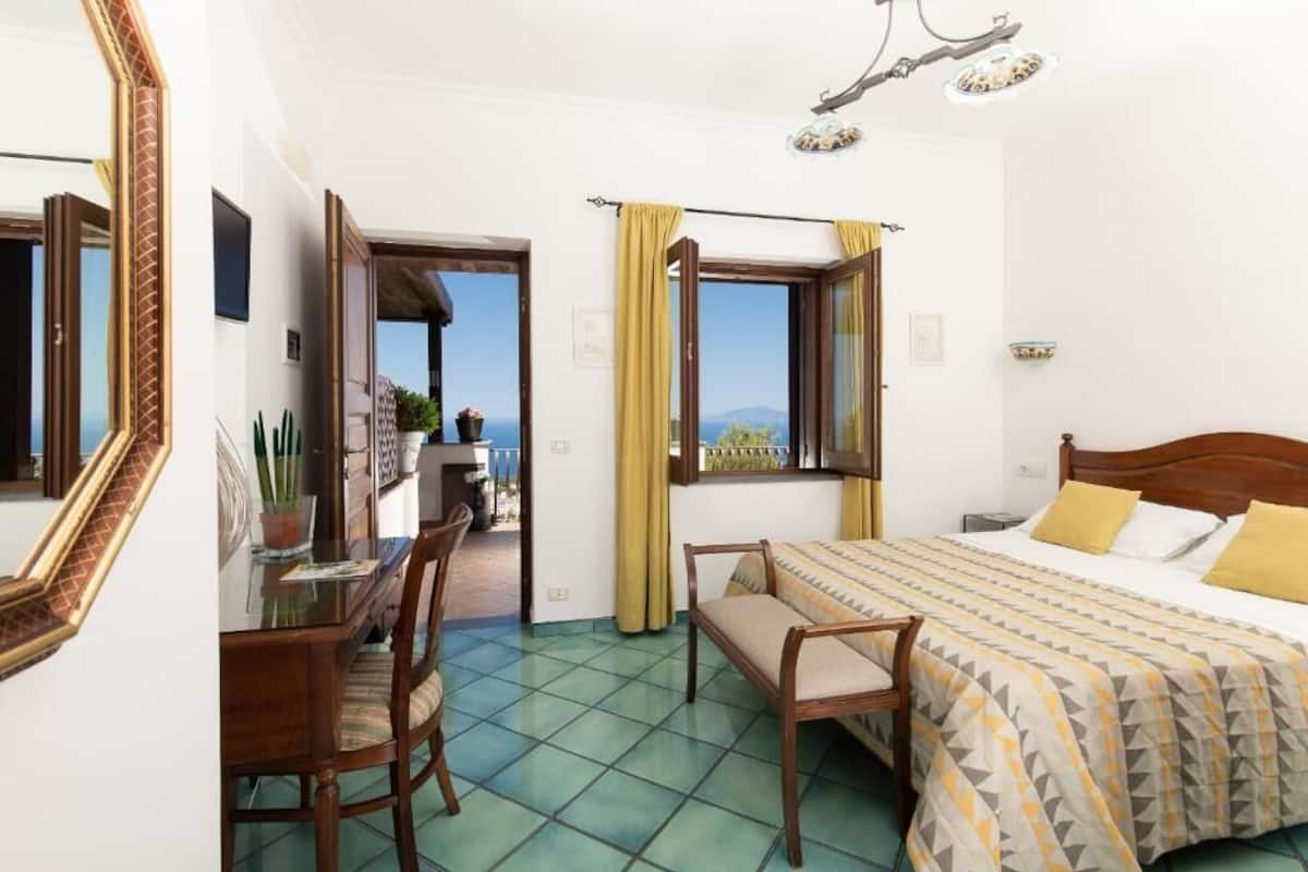 A hotel room with a bed, a desk, and a chair. There is a window with curtains, and a view of the outside.