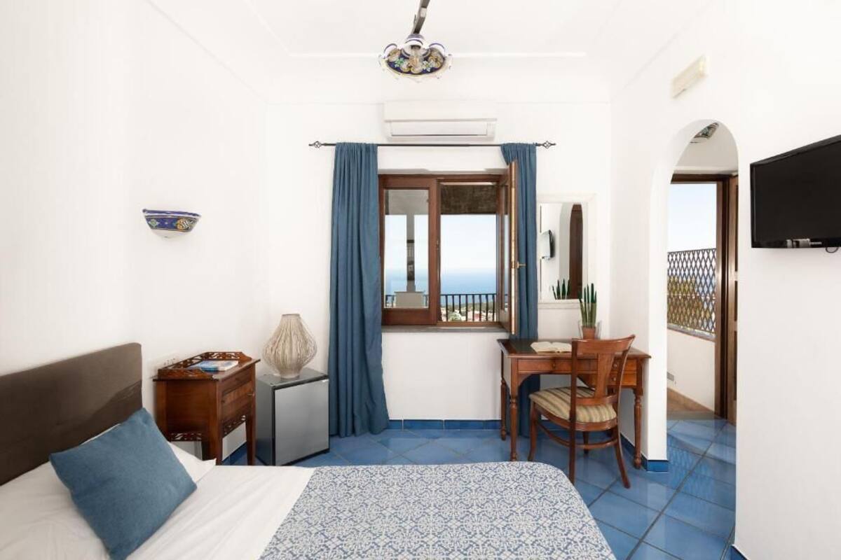 A bedroom with a bed, a desk, a chair, a TV, and a balcony with a view.