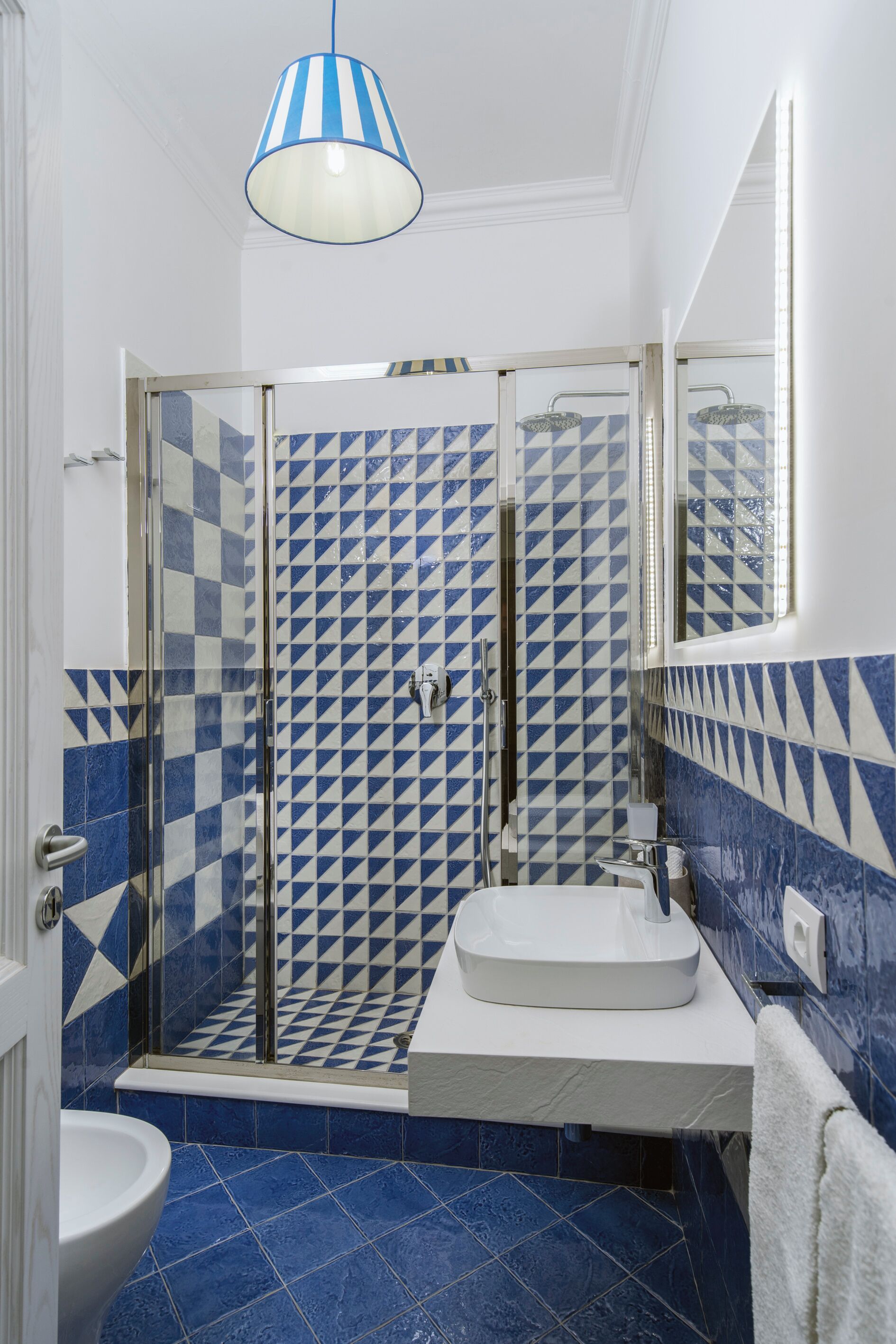 Un bany amb un disseny de rajoles geomètric, una cabina de dutxa de vidre, un lavabo blanc i una làmpada penjant.