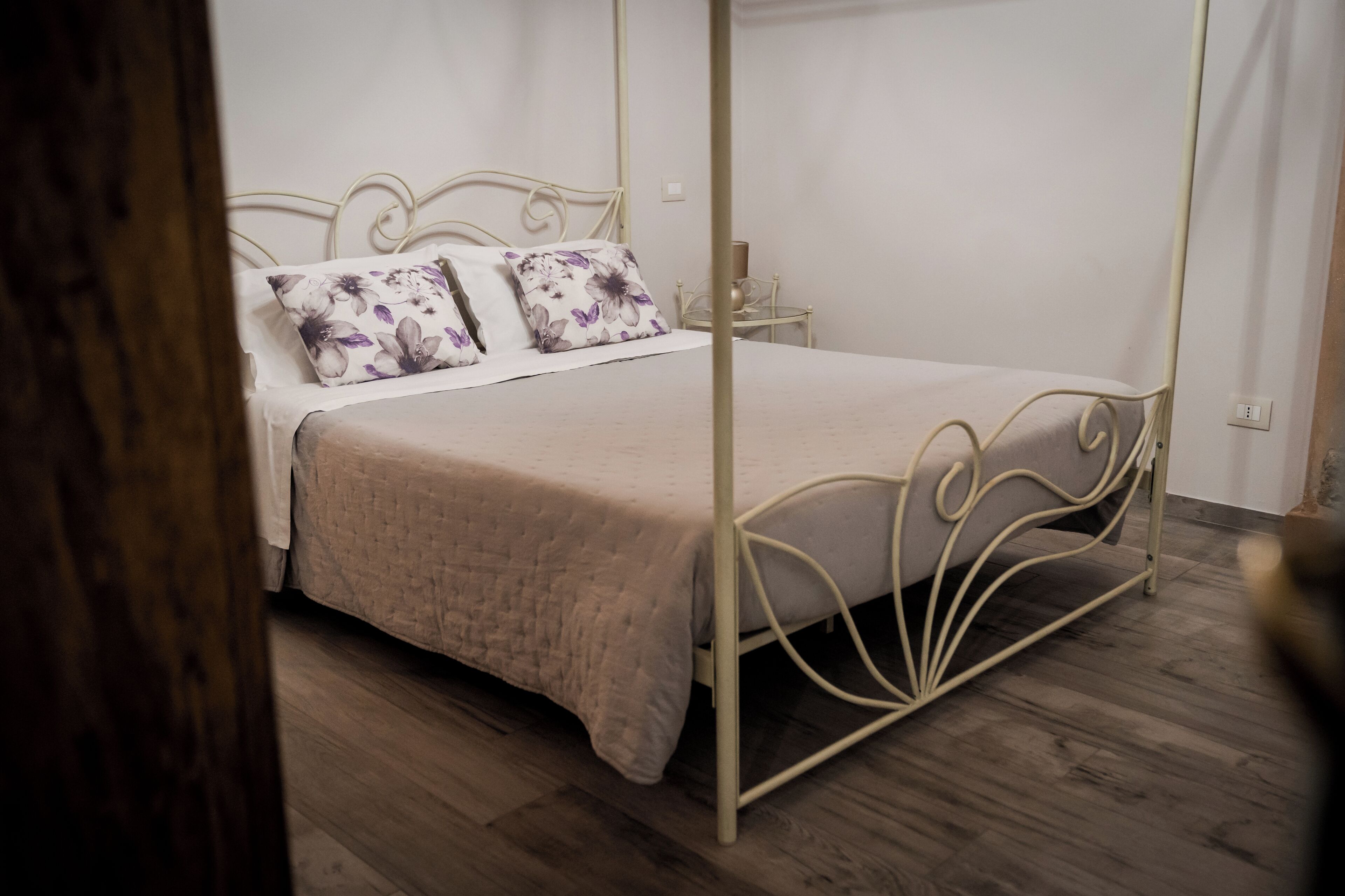 A four-poster bed with a beige bedspread and floral pillows, a bedside table with a lamp, and a wooden floor.