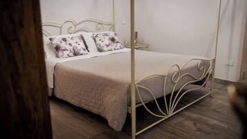 A four-poster bed with a beige bedspread and floral pillows, a bedside table with a lamp, and a wooden floor.