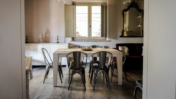 Un comedor con una mesa de madera, sillas de metal, una ventana con contraventanas abiertas y un espejo en la pared.