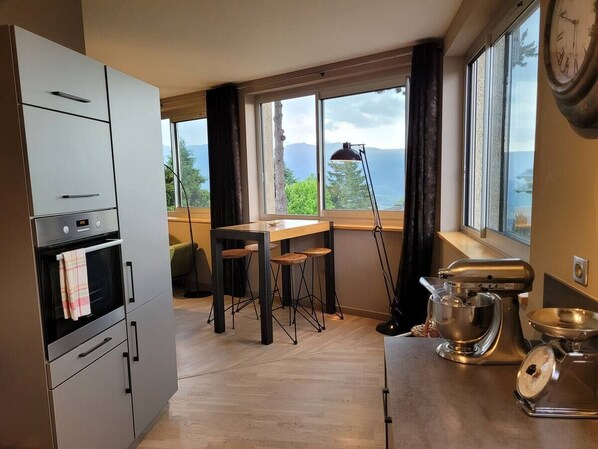A kitchen with a built-in oven, a dining table with stools, a large window with a view, and a stand mixer on the counter. - Cocon 1800, Downtown (Font-Romeu-Odeillo-Via)