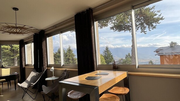 A spacious room with a wooden dining table, chairs, and large windows offering a view of trees and mountains.