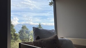 A bedroom with a bed against a wall, a window with a view of trees and mountains, and a pillow with a cat design.