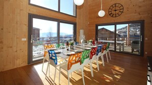 A dining area with a long table set for six, colorful chairs, large windows, and a wooden interior. - Chalet Archi, the mountain par excellence (Font-Romeu-Odeillo-Via)