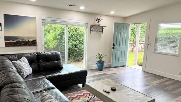 A modern living room with a black leather sofa, a coffee table, and a large window with a view of greenery. - The Bluff House~Upgraded & Close to Town (McKinleyville)