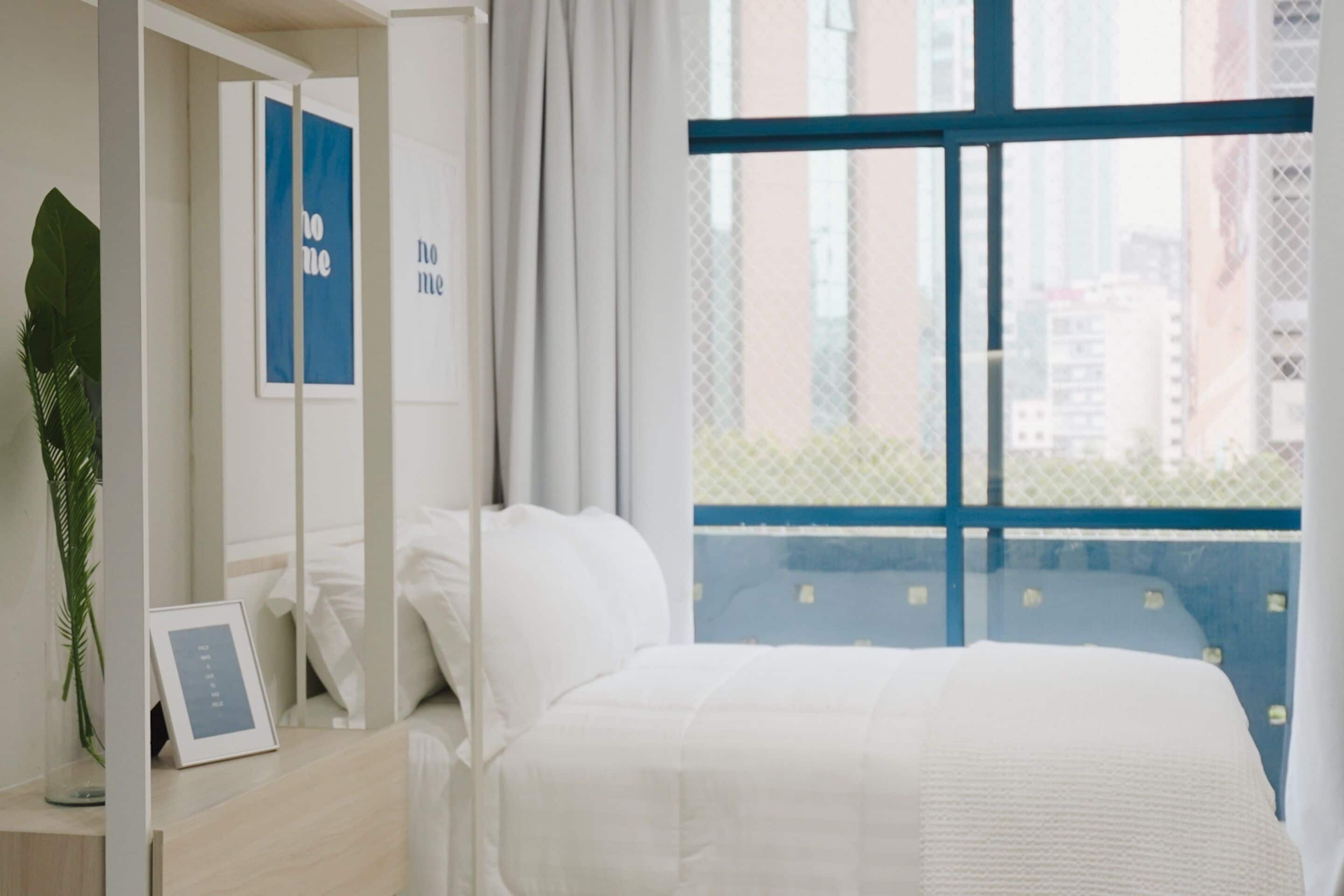 A hotel room with a large window, a bed with white bedding, a bedside table with a framed picture, and a potted plant.