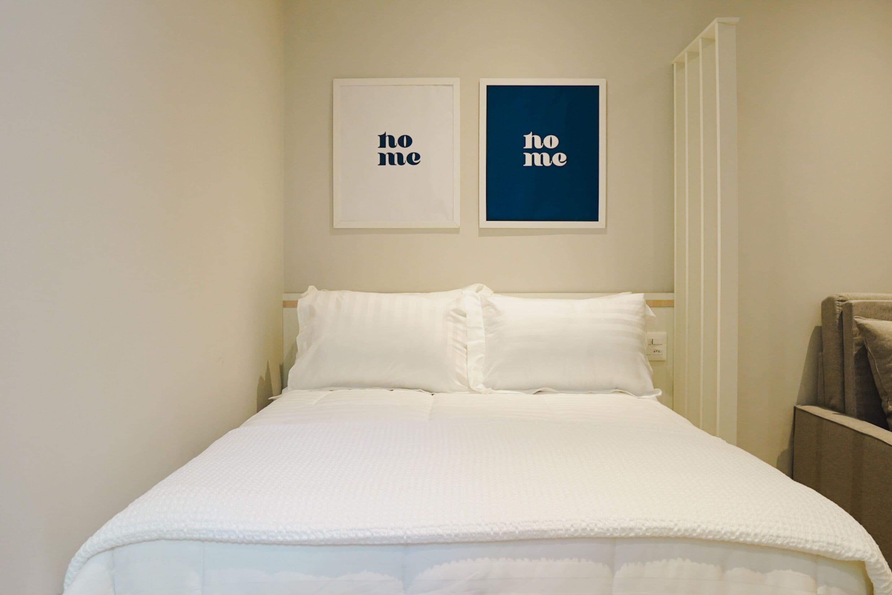 Uma cama bem arrumada com roupa de cama e almofadas brancas, duas placas na parede com a inscrição "no me", e uma porta branca à direita.