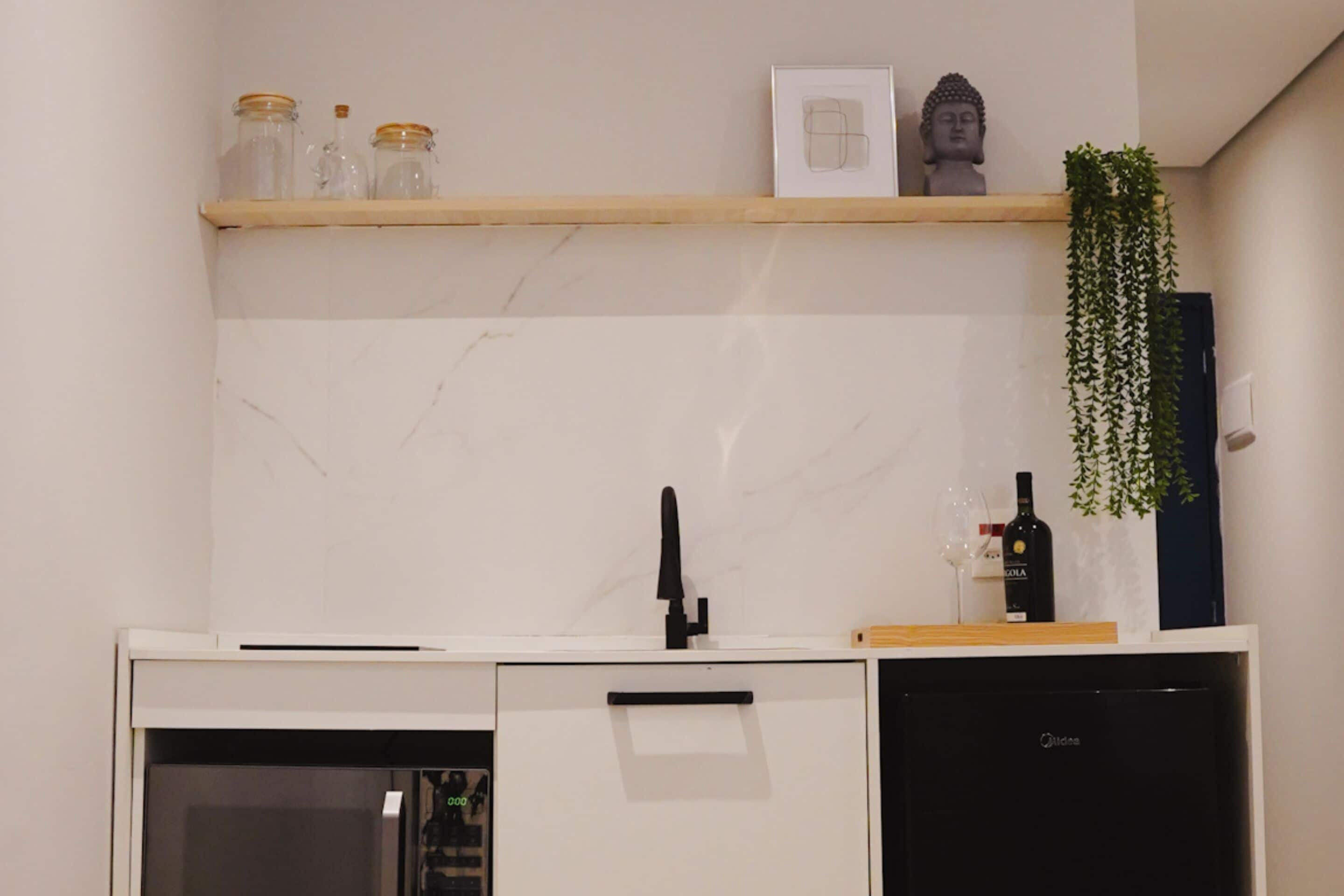 Una cocina con encimera blanca, pileta negra, refrigerador negro y una repisa de madera con frascos y una estatua de Buda.