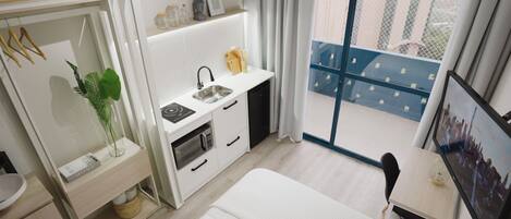A compact hotel room with a bed, a small kitchenette, a bathroom, and a balcony with a city view.