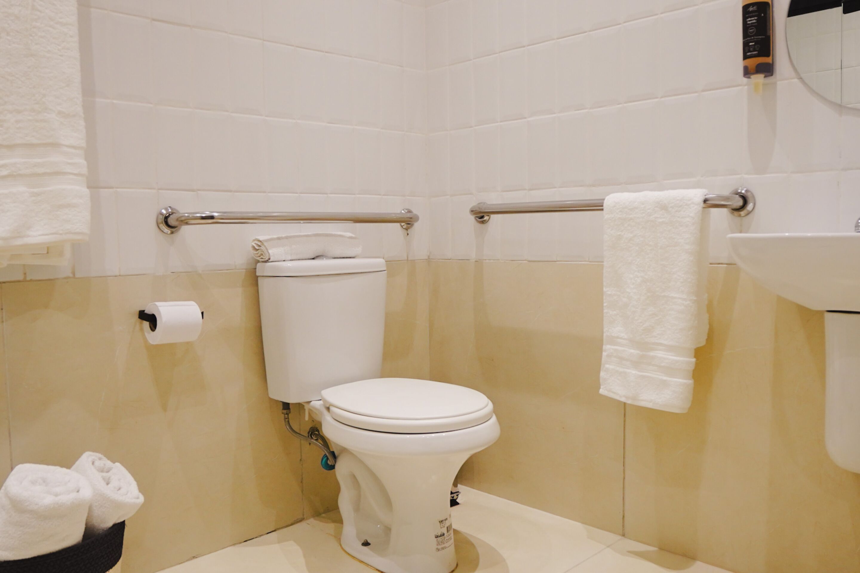 Un baño pequeño y limpio con inodoro, barras de apoyo, lavamanos y un perchero para toallas.