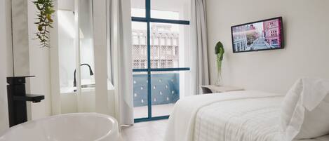 Quarto de hotel moderno com uma banheira grande e branca, cama com roupa de cama branca, televisão na parede e varanda com vista para edifícios.