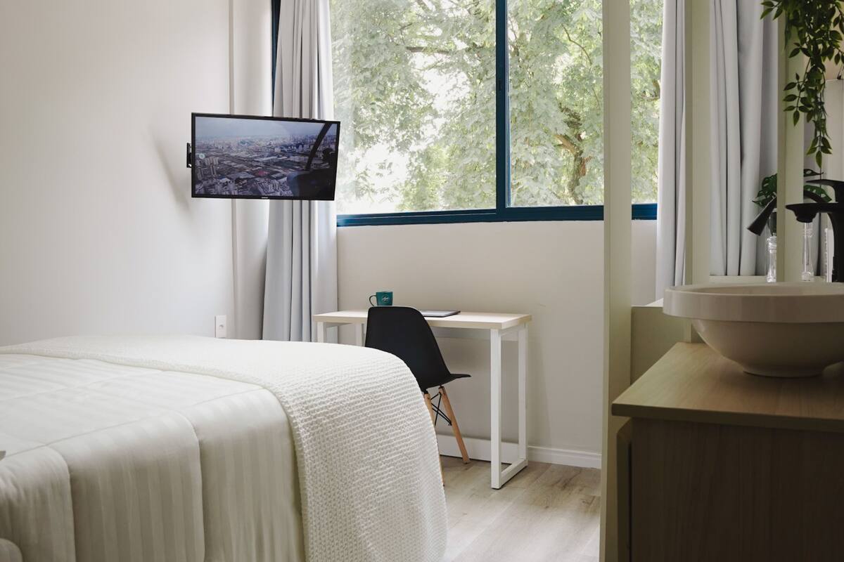 Ein Schlafzimmer mit Bett, Schreibtisch, Fernseher und Fenster mit Blick auf Bäume.
