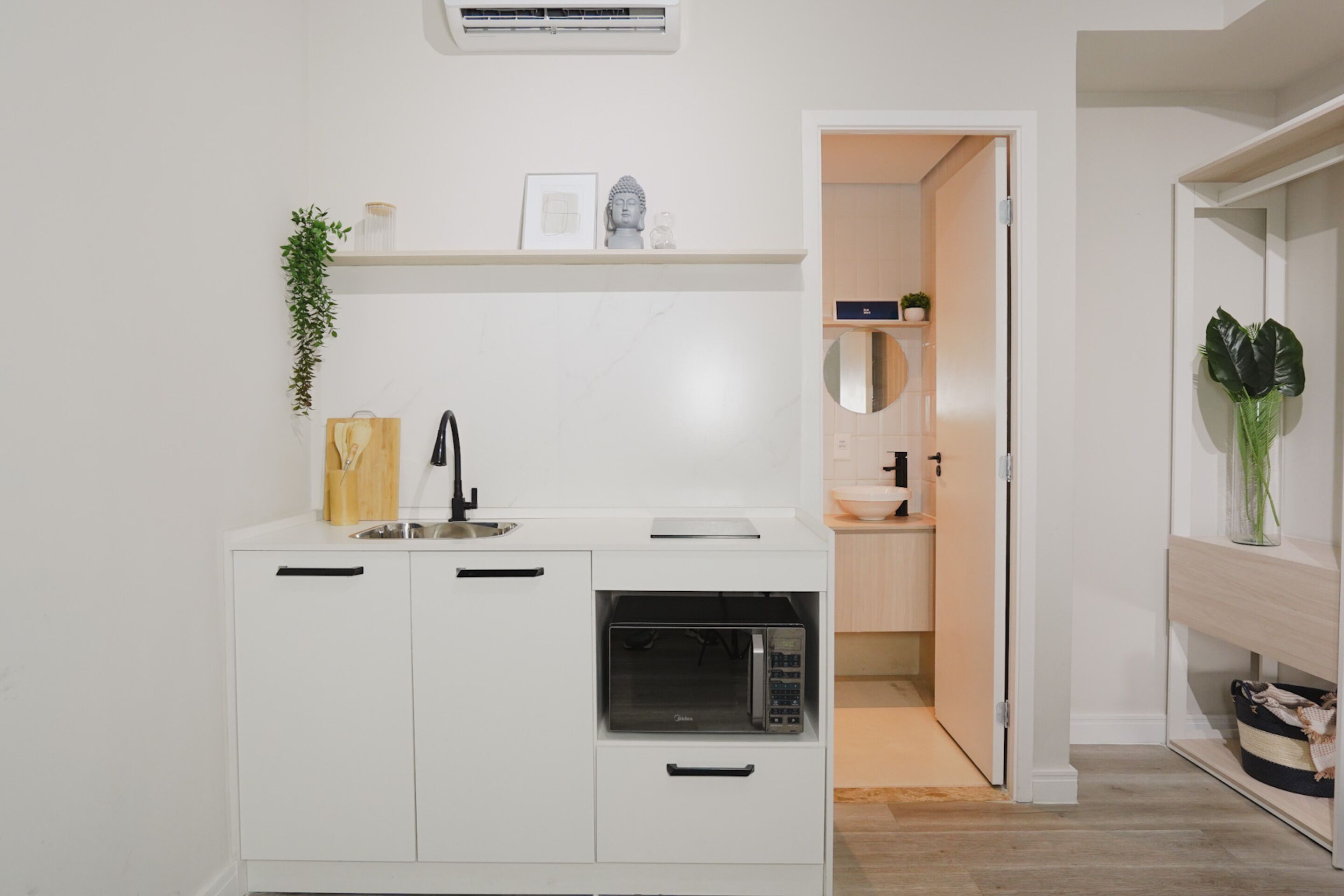 A compact kitchen with white cabinetry, a microwave, and a sink. There is a shelf above the sink with decorative items and a plant.