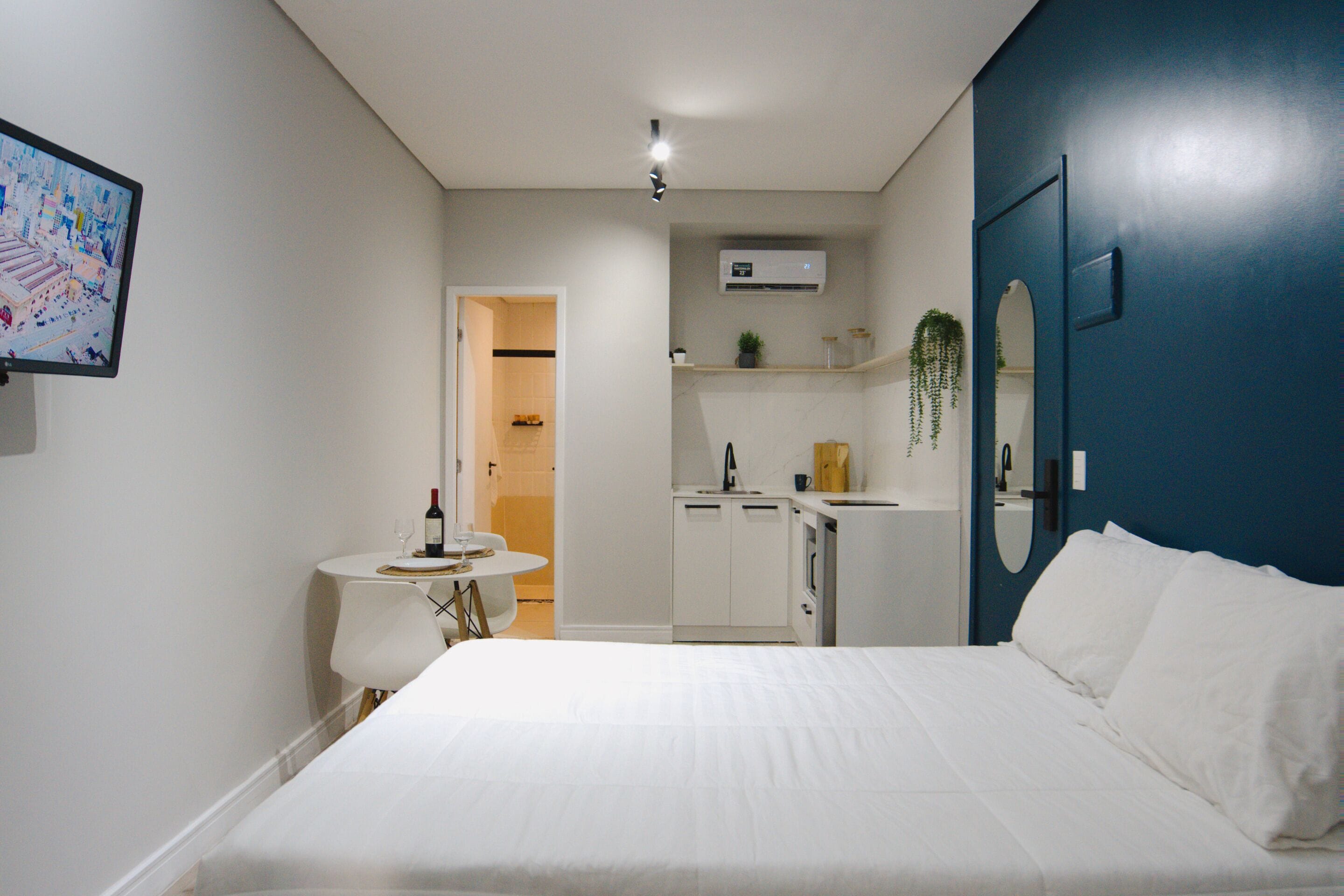 A hotel room with a bed, a small table with a wine bottle, a chair, a kitchenette, and a bathroom visible through an open door.