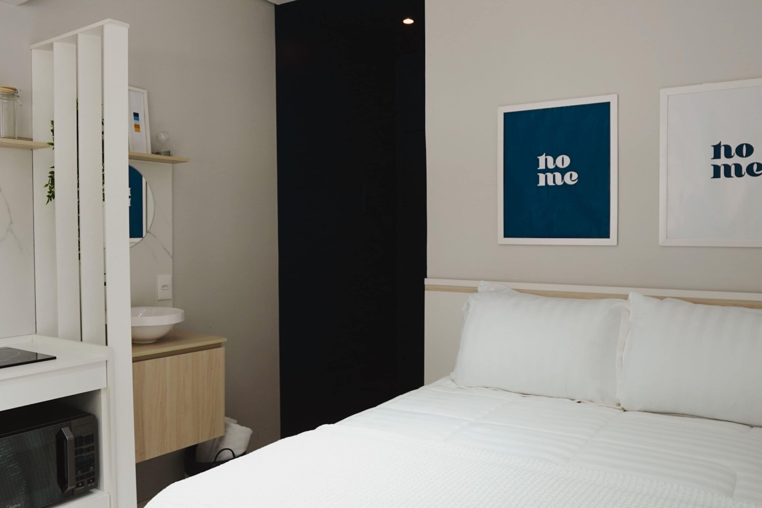 Una moderna camera d'albergo con un letto, biancheria da letto bianca, un comodino, un forno a microonde, un lavandino e opere d'arte incorniciate appese al muro.