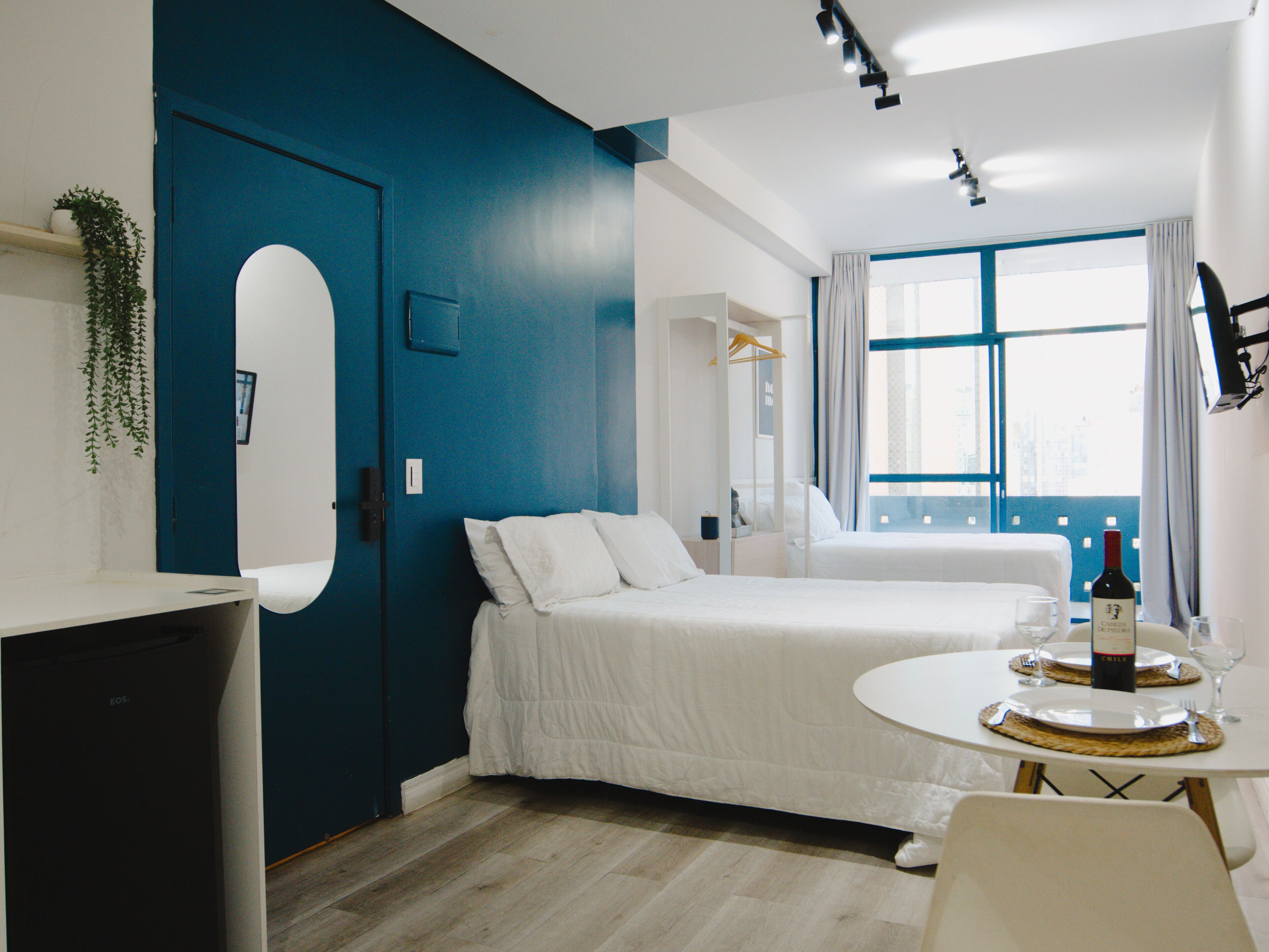 A hotel room with a bed, a desk, a chair, a window with curtains, and a bottle of wine on the table.