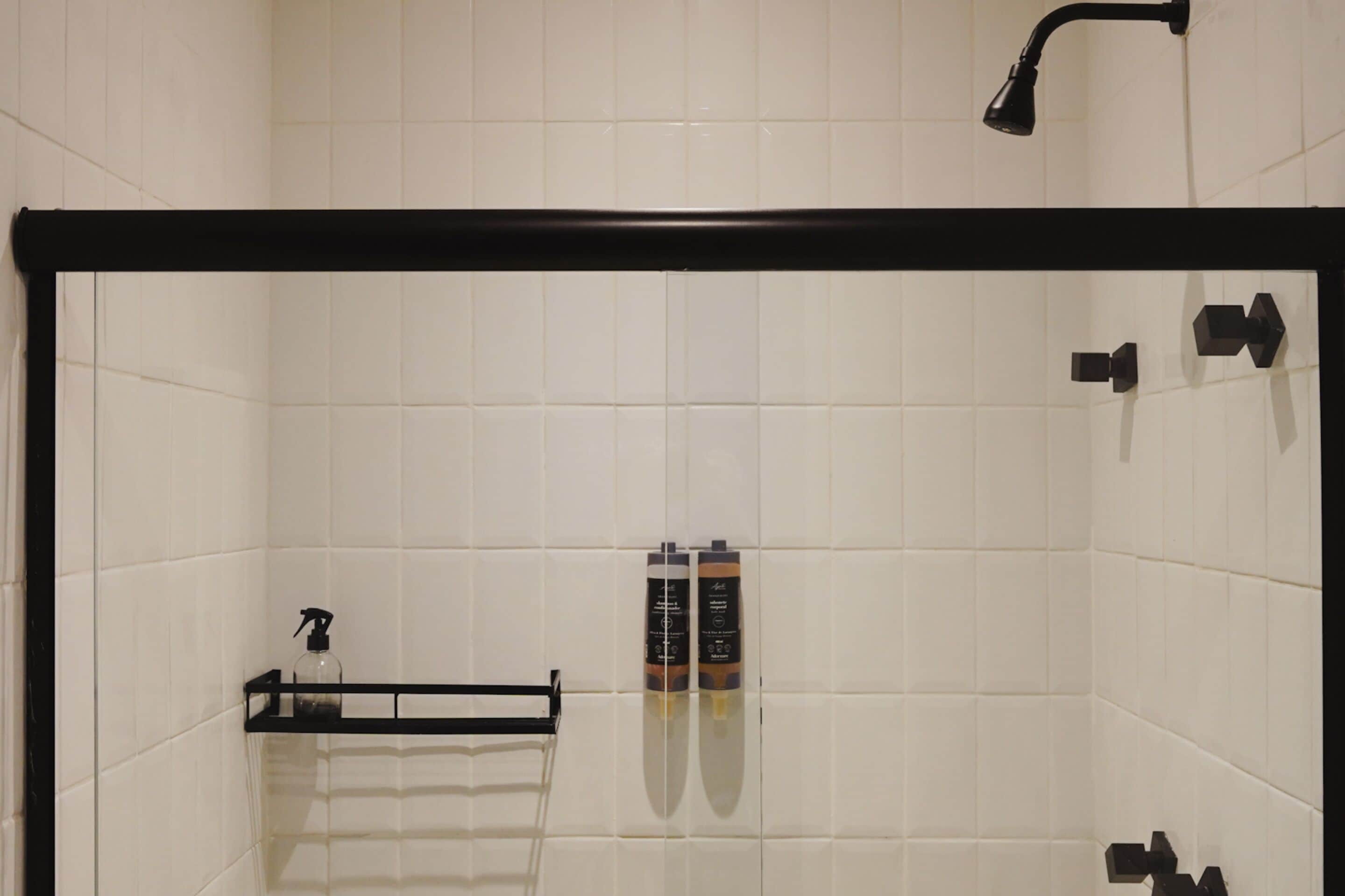 A bathroom with white tiled walls, a black shower enclosure, a glass shelf with toiletries, and a handheld showerhead.