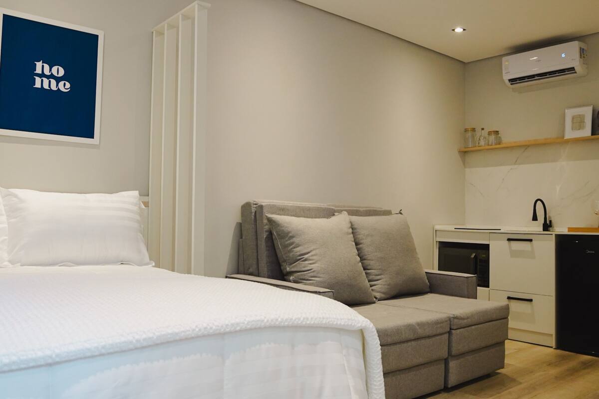 Ein modernes Schlafzimmer mit einem Bett, einem grauen Sofa und einer kleinen Küchenzeile.