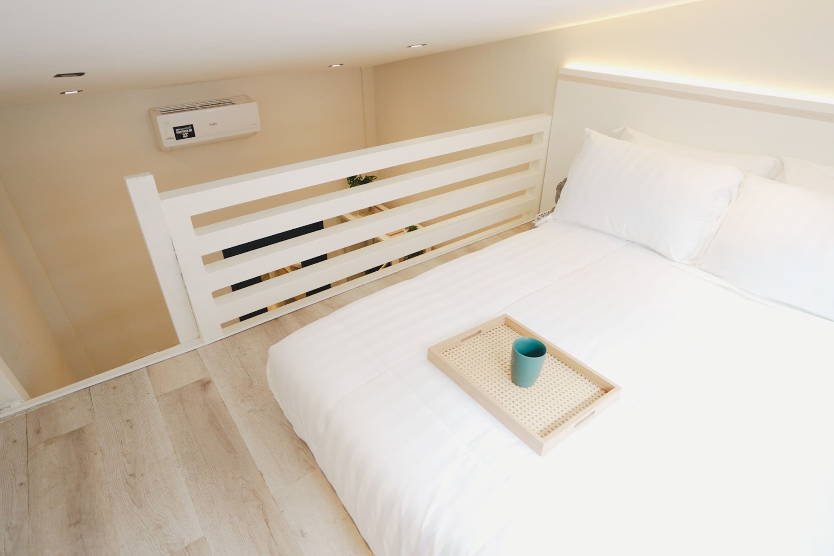 A bedroom with a white bed, a wooden headboard, a bedside table with a tray, and an air conditioning unit on the wall.