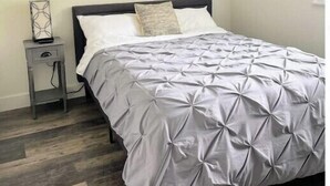 Un lit bien fait, recouvert d’une couverture grise, une petite table de chevet avec une lampe et un mur sobre.
