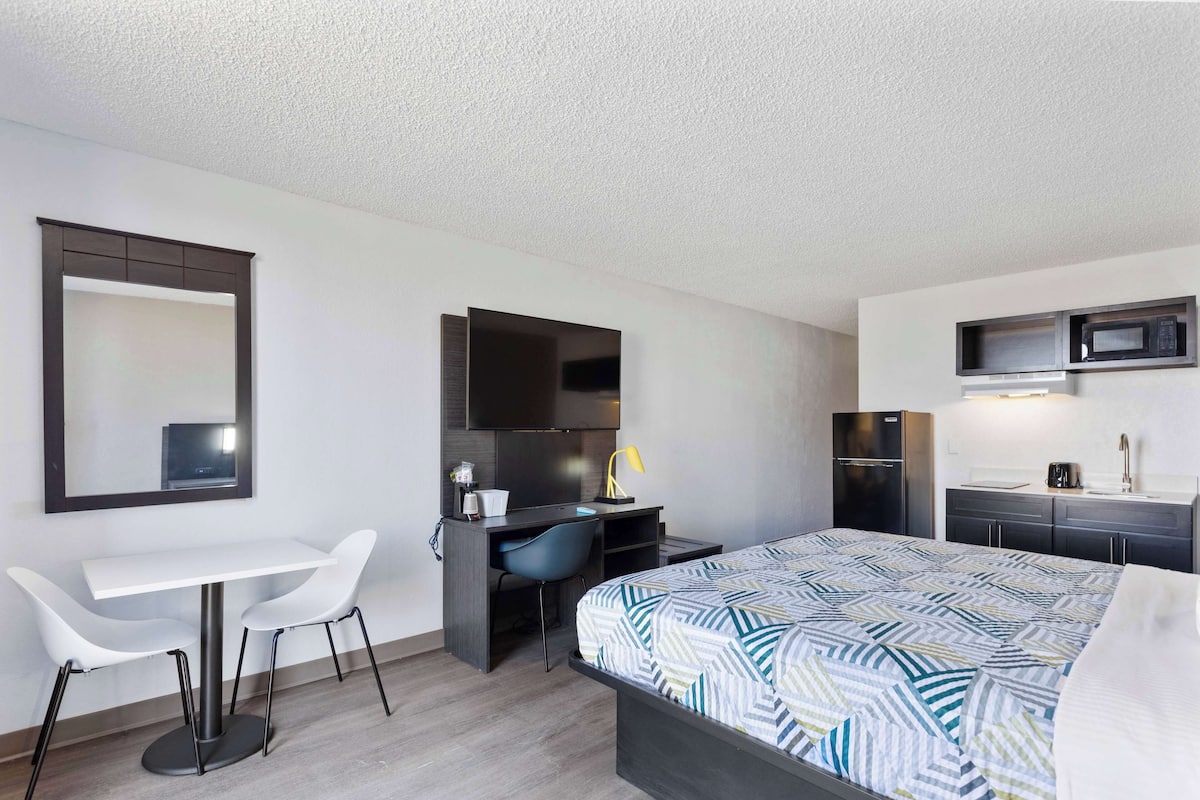 A modern hotel room with a bed, a desk with two chairs, a microwave, a refrigerator, and a small kitchenette.