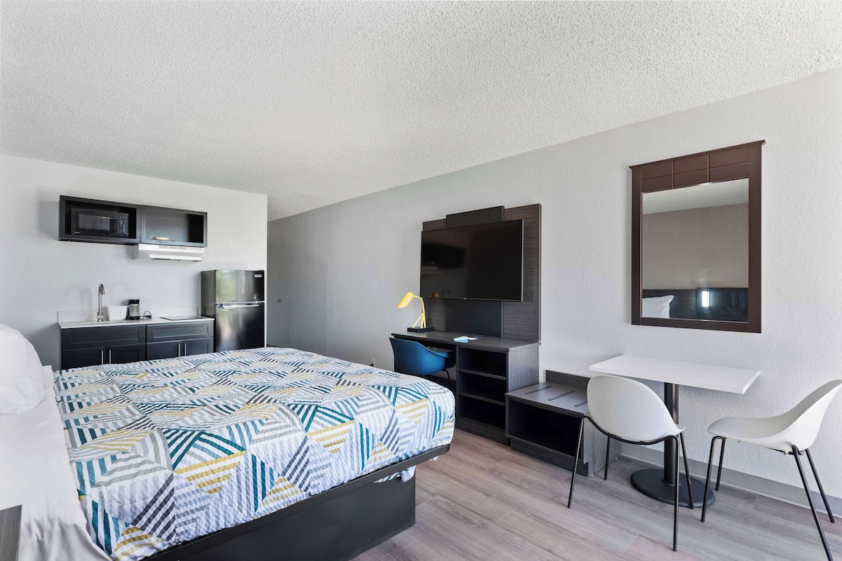 A modern hotel room with a bed, a desk, a chair, a TV, a mirror, and a kitchenette.