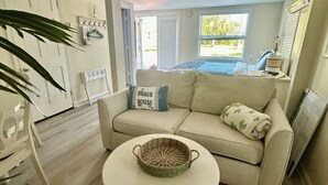 A modern living room with a beige sofa, a round coffee table with a woven centerpiece, and a large window providing a view of the outdoors. - FLAMINGO COTTAGE summer fun. Pool, boating, fishing lobster season.  (Summerland Key)