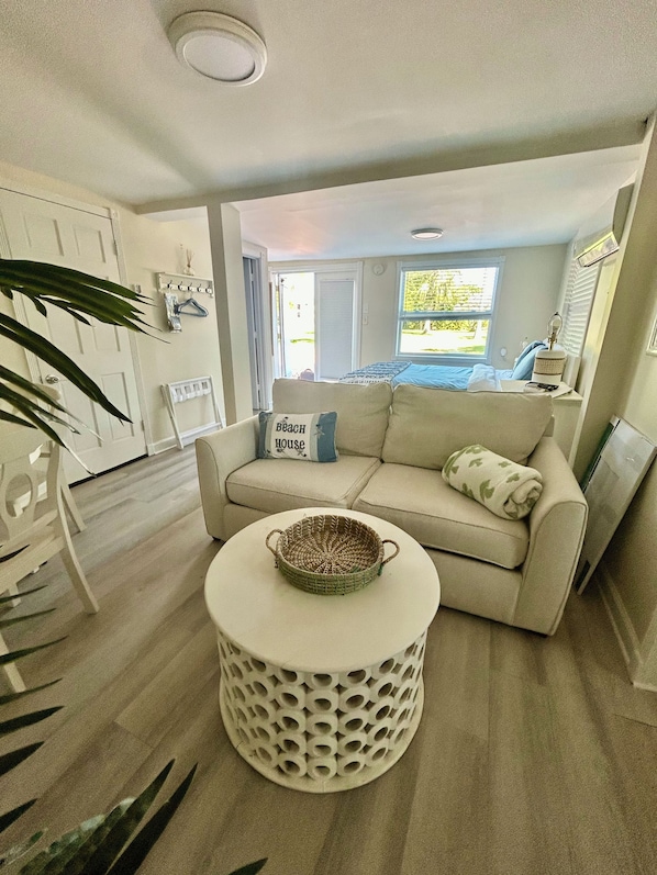 A modern living room with a beige sofa, a round coffee table with a woven centerpiece, and a large window providing a view of the outdoors. - FLAMINGO COTTAGE - Sleeps 2 adults and 2 children (Summerland Key)