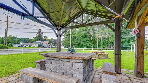 Un espace extérieur aménagé, abrité et doté de bancs en pierre et d’une table centrale, entouré de verdure et délimité par une clôture blanche.