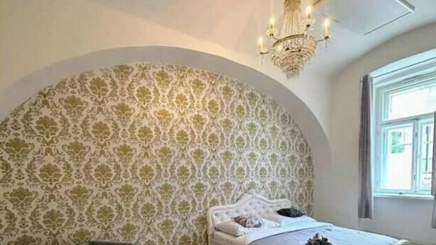 Une chambre à coucher avec une grande tête de lit en arc de cercle, un lit recouvert de linge de lit blanc, un canapé gris agrémenté de coussins à motifs et un lustre.