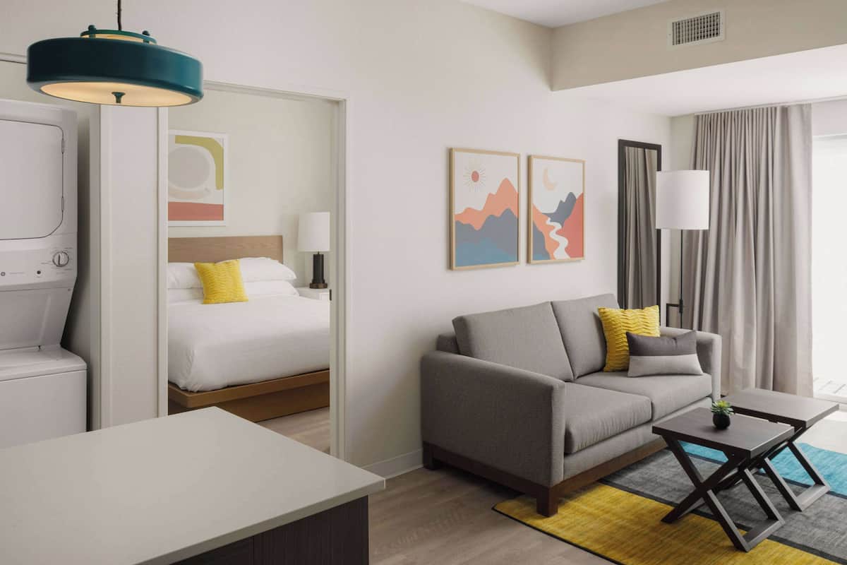 Ein modernes Hotelzimmer mit Bett, Sofa und einem kleinen Tisch. Ein Fenster mit Vorhängen, eine Wandleuchte und Kunstwerk an der Wand sind ebenfalls vorhanden.