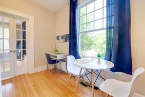 A room with a wooden floor, a white desk with a chair, a round table with a chair, and a window with blue curtains. - Historical 1BR Apt in Glenwood (Raleigh)
