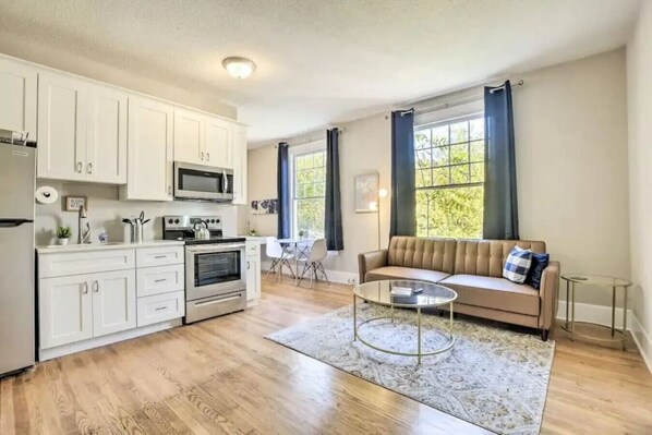 A modern kitchen with white cabinets, a microwave, and an oven. There is a beige sofa with a glass coffee table in the living area. - Historical 1BR Apt in Glenwood (Raleigh)