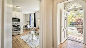 A modern kitchen with stainless steel appliances, a dining area with a glass table, and a view of a garden through a door. - Historical 1BR Apt in Glenwood (Raleigh)