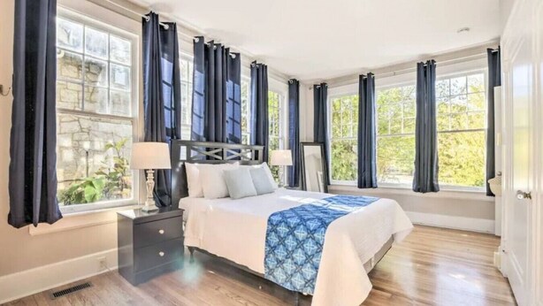 A spacious bedroom with a large bed, dark curtains, and a view of greenery outside. - Historical 1BR Apt in Glenwood (Raleigh)