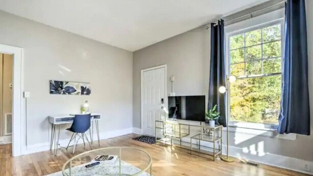 Un salon moderne avec un plancher en bois, une table basse en verre, un bureau avec une chaise, un téléviseur et une grande fenêtre avec des rideaux.