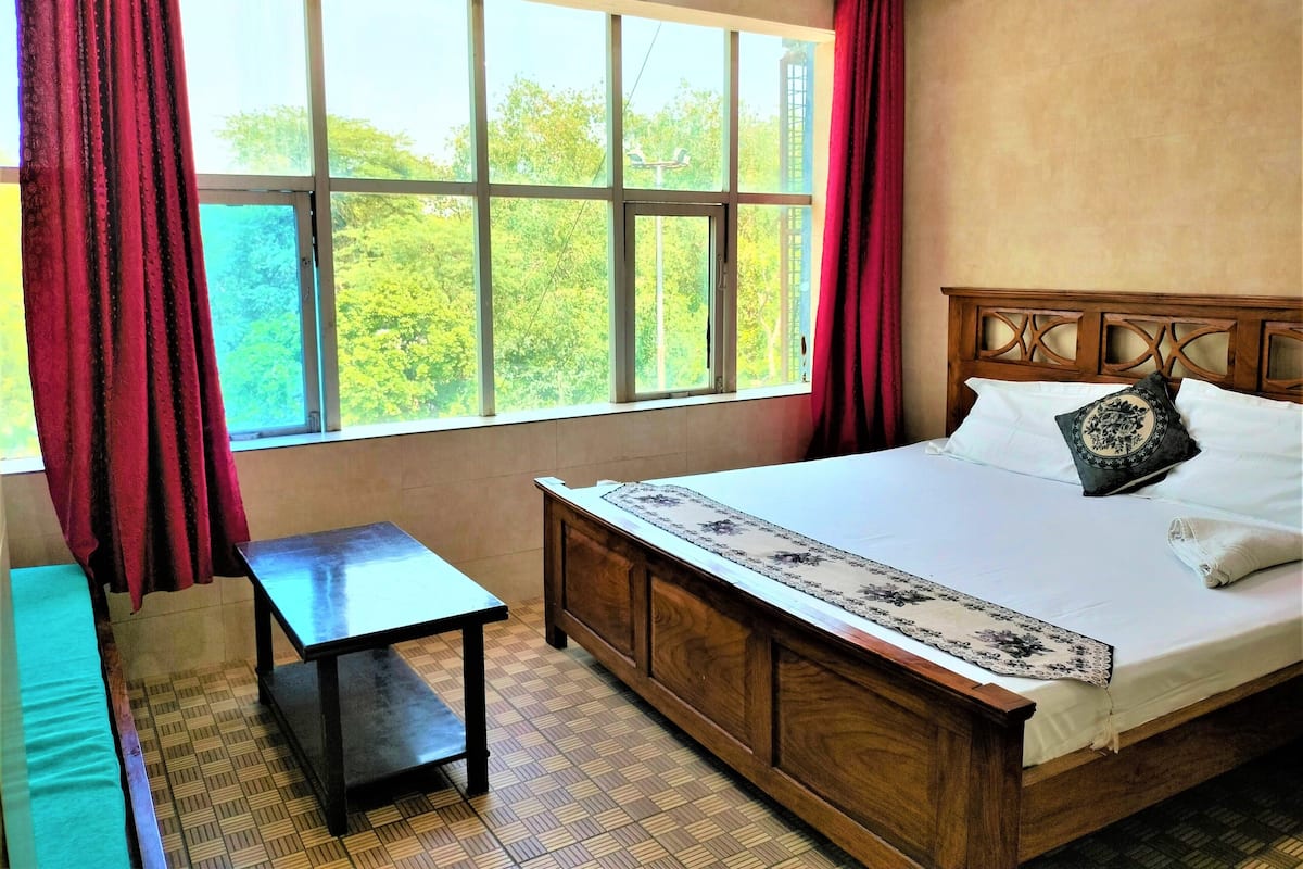 Une chambre d’hôtel avec un grand lit, une tête de lit en bois, une petite table et une vue sur la verdure par la fenêtre.