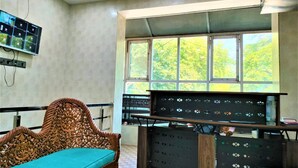 Ein Zimmer mit einem großen Fenster, einem Holzbstuhl mit türkisnem Kissen und einem Schreibtisch mit mehreren Bildschirmen.
