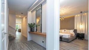 Ein modernes Schlafzimmer mit einem großen Bett, Nachttischen und einem Spiegel. Ein mit Holzboden ausgelegter Flur führt zu einer Treppe.
