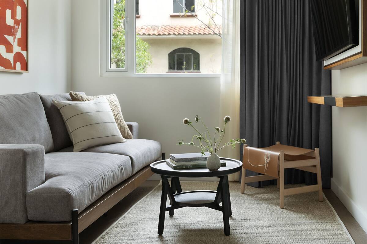 A modern living room with a grey sofa, a small round table with a vase, and a view of a building through a window.