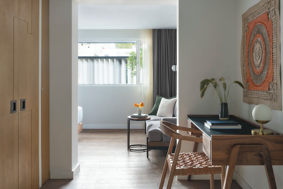 A modern, well-lit room with a wooden wardrobe, a small round table with a vase of flowers, a sofa with a green cushion, and a desk with a lamp and books.